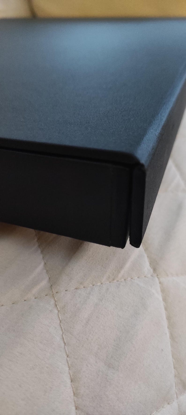 Peut inclure: Gros plan sur une bo&icirc;te noire &agrave; surface textur&eacute;e. La bo&icirc;te, avec un couvercle et une base, pr&eacute;sente une couture visible. Elle repose sur un tissu matelass&eacute; blanc cass&eacute;.