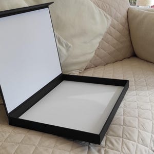 Peut inclure: Une bo&icirc;te de pr&eacute;sentation noire ouverte, r&eacute;v&eacute;lant un int&eacute;rieur blanc. La bo&icirc;te est rectangulaire et poss&egrave;de un couvercle articul&eacute;. Elle est pos&eacute;e sur une surface beige matelass&eacute;e, avec des coussins beiges en arri&egrave;re-plan.