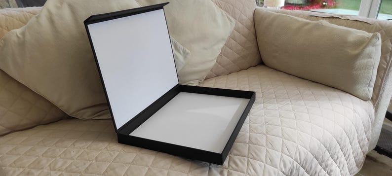 Peut inclure: Une bo&icirc;te de pr&eacute;sentation noire ouverte, r&eacute;v&eacute;lant un int&eacute;rieur blanc. La bo&icirc;te est rectangulaire et poss&egrave;de un couvercle articul&eacute;. Elle est pos&eacute;e sur une surface beige matelass&eacute;e, avec des coussins beiges en arri&egrave;re-plan.