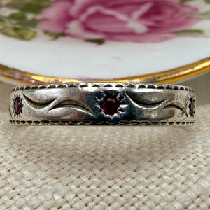 May include: A silver ring featuring a repeating leaf design and set with red gemstones. The ring is displayed against a floral patterned plate with a gold rim, and a textured beige surface.