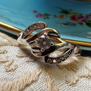 May include: A silver-toned ring featuring a central round clear stone set within a swirling, open-work band. The band is accented with small, embedded clear stones along the curved metal segments that wrap around the centre setting.