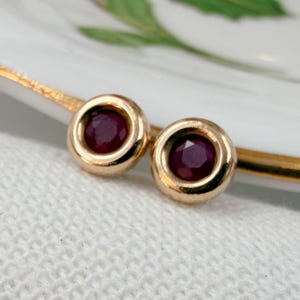 May include: A pair of round stud earrings featuring deep red circular gemstones set in polished gold-toned metal bezels. The earrings have a smooth, minimalist design with a faceted surface on the stones that reflects light.