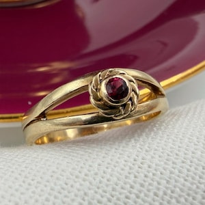 May include: A gold ring featuring a central red gemstone, encircled by a textured, decorative setting. The ring's band splits into two parallel sections. The ring is presented on a white surface, with a burgundy and gold-edged saucer in the background.