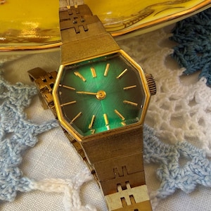 May include: A gold-tone wristwatch featuring an octagonal case and a vibrant emerald green sunburst dial with gold-tone hour markers and hands. The watch includes a matching gold-tone metal link bracelet with a textured, brushed finish.