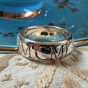 May include: A silver-toned metal ring featuring the Roman numerals MCMII engraved around the outer band. The interior of the band displays a small hallmark stamp. The ring has a polished finish and a wide, uniform band construction.