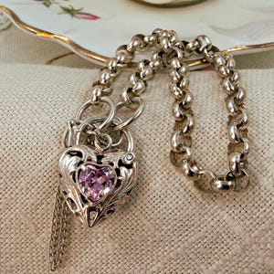 May include: A silver necklace with a heart-shaped pendant. The pendant has a pink gemstone and detailed design. The necklace is shown on a neutral surface, with a floral teacup visible in the background.