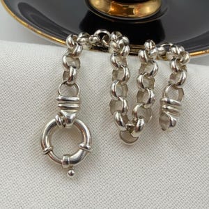 May include: A silver chain necklace featuring round, interlocking links and a large, circular spring-ring clasp. The metal has a polished, reflective finish, and the design consists of a continuous series of uniform, thick, rounded metal loops.