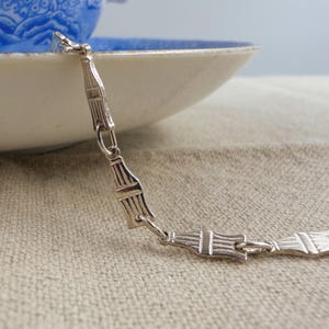 May include: A silver-toned metal chain bracelet featuring a series of repeating, elongated, rectangular links with vertical grooved detailing. The chain is composed of individual segments connected by small circular jump rings, creating a uniform pattern.