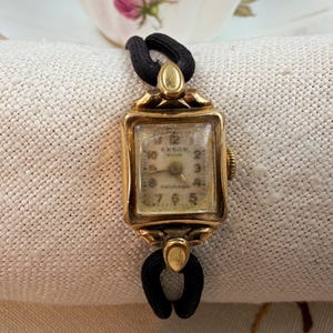 May include: A vintage gold wristwatch with a rectangular face and a black elastic strap. The watch face displays the numbers 1 to 12 and the brand name "Orson". The watch is set against a neutral-toned background.