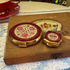 May include: Two round and hexagonal gold-tone metal compact mirrors featuring a deep burgundy border and a central cream-coloured inlay decorated with pink floral patterns. The compacts are resting on a wooden box with a vintage label.