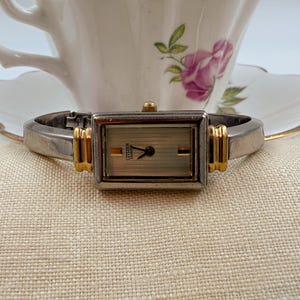 May include: A Citizen Quartz watch with a rectangular face and a silver and gold strap. The watch face is a light champagne colour with black hands and the Citizen Quartz logo. The watch is displayed on a beige surface.