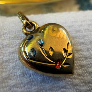 May include: A gold-toned heart-shaped pendant featuring an engraved floral motif with small blue and red accents. The word MUM is etched into the upper right section of the heart. A circular metal bail is attached to the top for hanging.
