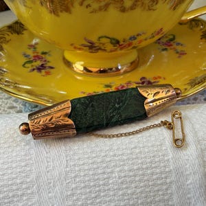 May include: A bar brooch featuring a rectangular dark green stone centre with gold-toned metal end caps engraved with leaf patterns. The brooch includes a small gold-toned safety pin attached by a thin metal chain to the main bar structure.