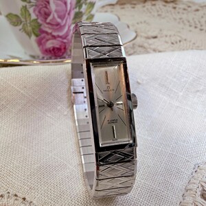 May include: A vintage Milus wristwatch with a silver rectangular face and a silver expanding band. The watch face displays the brand name and "17 Jewels Incabloc" text. The watch is Swiss made.