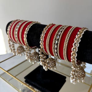 May include: Red bangles with gold and pearl embellishments, presented on a black stand. The bangles have detailed patterns and hanging gold-coloured bells with pearl drops. The jewellery is displayed on a mirrored surface.