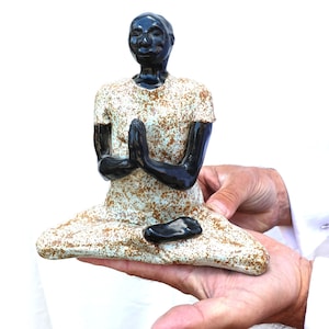 Könnte beinhalten: Eine Keramikskulptur einer Person in einer sitzenden Yoga-Pose. Die Figur hat schwarze Haut und trägt ein helles Oberteil mit braunen Sprenkeln. Die Hände sind in Gebetshaltung gefaltet. Die Skulptur wird in den Händen einer Person gehalten.