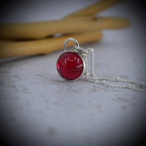 May include: A silver necklace with a circular, deep red pendant. The pendant is encased in a silver setting and has a glossy finish. The chain is fine, with tiny silver beads spaced along its length.