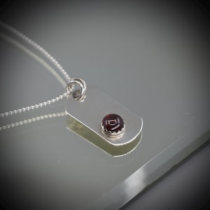 May include: A silver pendant necklace. The pendant is a rectangular tag with a small, round, dark red gemstone. The pendant hangs from a delicate silver chain. The gemstone is set in a silver bezel.