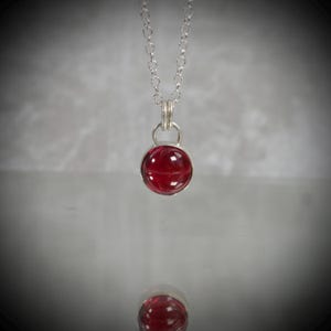 May include: A silver necklace featuring a round, deep red cabochon gemstone set in a simple silver bezel. The pendant hangs from a delicate silver chain, with the circular red stone catching the light and reflecting on the surface below.