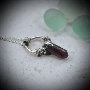 May include: A silver necklace featuring a dark red, hexagonal, pointed gemstone pendant suspended from a circular silver ring. The ring is accented with small, textured silver beads on either side of the stone, attached to a delicate silver chain.