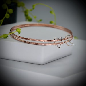 May include: Three copper bangles with a hammered finish, one with a silver star charm and another with a crescent moon charm. The bracelets are displayed on a white surface. The bangles are a beautiful piece of jewellery.