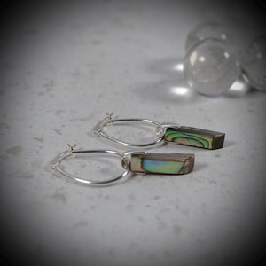 Könnte beinhalten: Ein Paar silberne Creolen mit baumelnden, rechteckigen Abalone-Muschel-Anhängern. Die Muschelstücke zeigen schillernde Grün-, Blau- und Violetttöne und sind an dünnen, polierten Silberreifen mit einem sicheren Verschlussmechanismus befestigt.