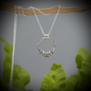 May include: A silver-colored necklace with a circular pendant. The pendant has a ring design with small decorative beads and a chain. The necklace is displayed against a gray background with green leaves.