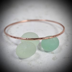 May include: A thin, hammered copper bangle bracelet positioned behind three smooth, rounded pieces of pale green sea glass. The metal band features a textured, reflective surface that contrasts with the matte finish of the sea glass stones.