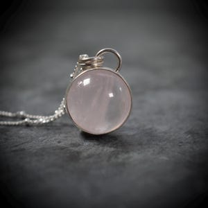 May include: A round, pale pink rose quartz gemstone pendant set in a silver-coloured metal bezel. The pendant features a small, coiled silver-coloured wire loop at the top, which is attached to a delicate, thin silver-coloured beaded chain necklace.