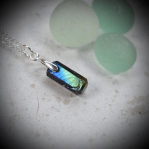 May include: A rectangular labradorite pendant featuring iridescent blue and green flashes, suspended from a delicate silver chain. The stone has a polished, faceted surface with visible internal fractures and a metallic silver bail attachment.