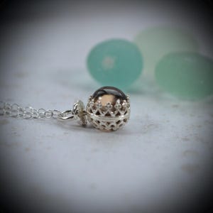 May include: A silver necklace featuring a spherical pendant encased in a decorative, crown-like filigree cage. The pendant holds a smooth, dark brown, translucent stone bead. The chain is composed of delicate, interlocking silver circular links.
