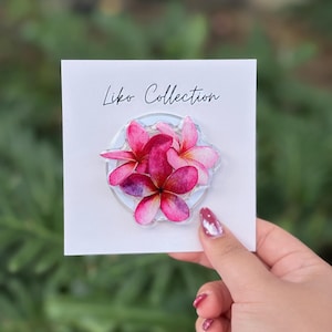 May include: A clear phone grip featuring a cluster of pink and magenta plumeria flowers. The flowers are arranged on a clear circular base, attached to a white card with the text "Liko Collection" in black script.