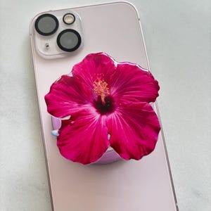 May include: A pink smartphone with a vibrant pink hibiscus flower phone grip attached. The flower has a detailed center and petals, and the phone has a clear camera lens protector.