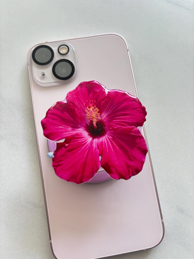 May include: A pink smartphone with a vibrant pink hibiscus flower phone grip attached. The flower has a detailed center and petals, and the phone has a clear camera lens protector.