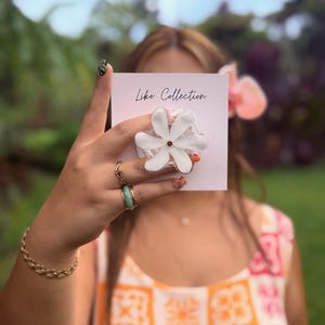 May include: A white flower-shaped phone grip with a central white petal and a clear, textured base. The grip is held in front of a person, obscuring their face. The card behind the grip reads "Liko Collection".