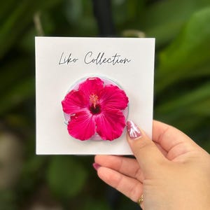 May include: A vibrant pink hibiscus flower phone grip on a white card. The card reads "Liko Collection" in script. The phone grip is held against a blurred green background.