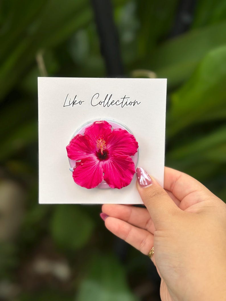 May include: A vibrant pink hibiscus flower phone grip on a white card. The card reads "Liko Collection" in script. The phone grip is held against a blurred green background.