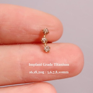 May include: A gold-colored, implant grade titanium earring with three small, clear gemstones in a vertical line. The text "Implant Grade Titanium" is visible, along with measurements of 16.18, 20g / 5.6, 7, 8, 10mm.