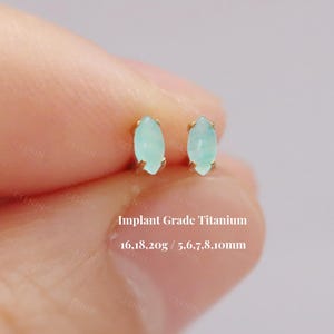 Pode incluir: Dois brincos pequenos em forma de marquesa verde claro com engastes dourados. O texto "Implant Grade Titanium" e os detalhes de tamanho são visíveis. Os brincos são exibidos em um fundo suave e desfocado.