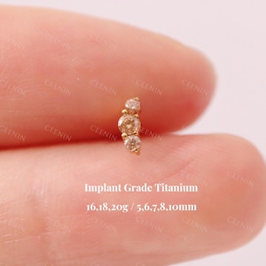 Könnte beinhalten: Ein goldfarbener, gebogener Piercing-Stecker mit drei klaren, runden Edelsteinen. Der Text "Implant Grade Titanium" und die Maße sind sichtbar. Der Schmuck wird zwischen den Fingern gehalten.