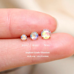 K&ouml;nnte beinhalten: Drei Gr&ouml;&szlig;en von Opal-Ohrringen in einer Hand. Die Ohrringe haben einen Durchmesser von 3 mm, 4 mm und 5 mm. Die Ohrringe sind aus implantatf&auml;higem Titan. Der Text "Implant Grade Titanium 16,18,20g / 5,6,7,8,10mm" befindet sich unter den Ohrringen.