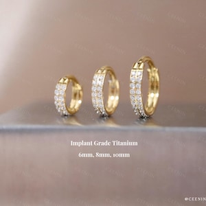 May include: Three gold-colored hoop earrings with embedded clear stones are displayed. The earrings are of varying sizes: 6mm, 8mm, and 10mm. The text "Implant Grade Titanium" is visible below the earrings.