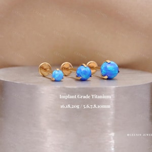 May include: Four gold-coloured stud earrings with blue opal-like spheres are displayed on a silver surface. The text "Implant Grade Titanium" is visible. The earrings vary in size.