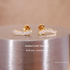May include: A pair of gold-coloured stud earrings with a curved design. Each earring features a row of small, clear gemstones. The text "Implant Grade Titanium" and measurements are visible.