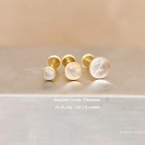 May include: Three gold-toned flat back stud earrings with round, translucent white stones. The text reads: Implant Grade Titanium, 16, 18, 20g / 5, 6, 7, 8, 10mm. The earrings are arranged in a row on a metallic surface.