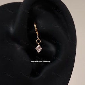 May include: A gold-colored earring with a small, clear, square-cut gemstone pendant. The earring is suspended from a curved hook and a small ring. The text "Implant Grade Titanium" is visible.