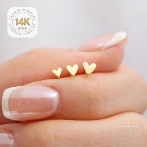 May include: Three gold heart-shaped stud earrings of varying sizes. The earrings are displayed on a person's finger. The earrings are made of 14K gold. The image includes a logo that says "CEENIN JEWELRY 14K GOLD".