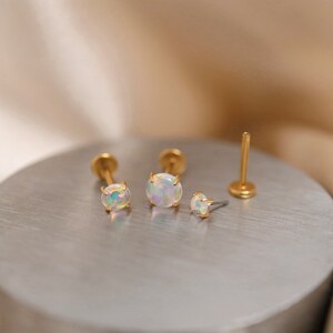 K&ouml;nnte beinhalten: Drei goldfarbene Opal-Ohrstecker mit einer Krappenfassung. Die Ohrringe liegen auf einer silbernen Oberfl&auml;che.