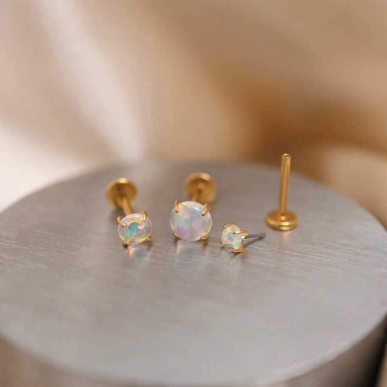 K&ouml;nnte beinhalten: Drei goldfarbene Opal-Ohrstecker mit einer Krappenfassung. Die Ohrringe liegen auf einer silbernen Oberfl&auml;che.