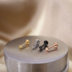 Puede incluir: Cuatro anillos de nariz en forma de estrella en diferentes colores: oro, plata, negro y oro rosa. Los anillos se muestran en una superficie plateada.